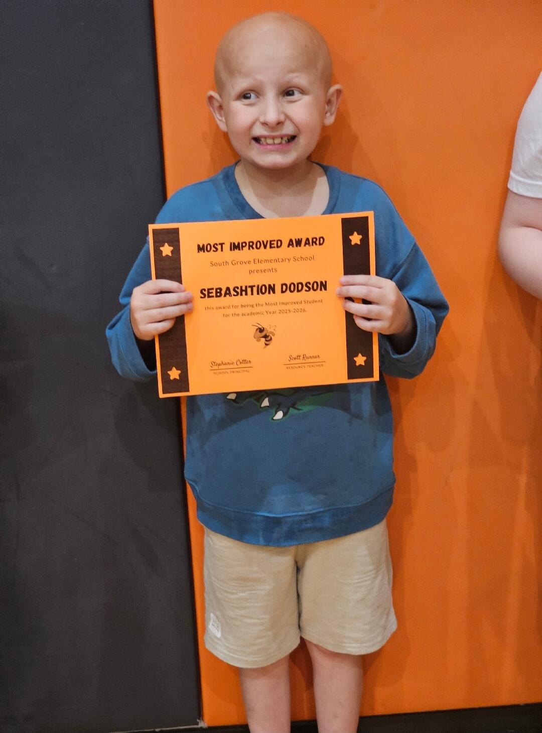 MOST IMPROVED AWARD. South Grove Elementary School presents SEBASHTION DODSON. This award is for being the Most Improved Student for the academic Year 2025-2026. Signed by Stephanie Cotter, School Principal, and Scott Runner, Resource Teacher. A young boy with a bald head is holding up an orange certificate, making a funny, happy face and smiling b