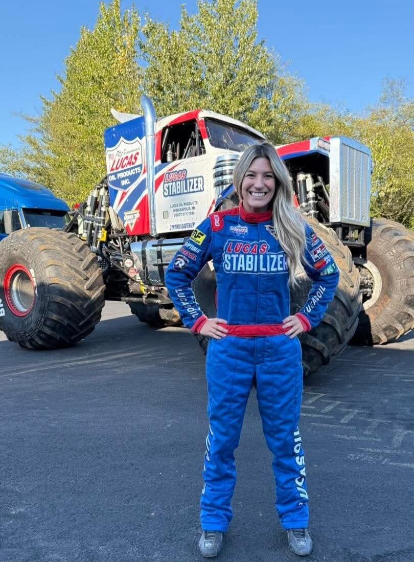 A woman in a blue racing suit with 'Lucas Stabilizer' and 'Lucas Oil' logos stands in front of a large monster truck. The truck also features 'Lucas Oil Products Inc.' and 'Lucas Stabilizer' branding, along with the name 'Cynthia Gauthier'.