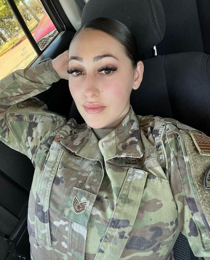 A female soldier in camouflage uniform sitting in a car, posing for a selfie.