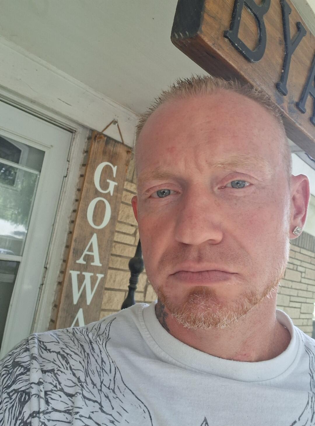 A man is taking a selfie. In the background, there are wooden signs with text 'G O A W' and 'BY' partially visible.