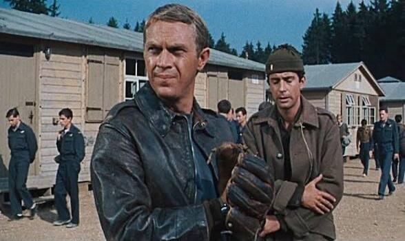 A scene from a classic war film, featuring two men, one holding a baseball glove, in what appears to be a prisoner-of-war camp. The man in the foreground with the leather jacket is Steve McQueen, and the man next to him in the military jacket is James Garner.