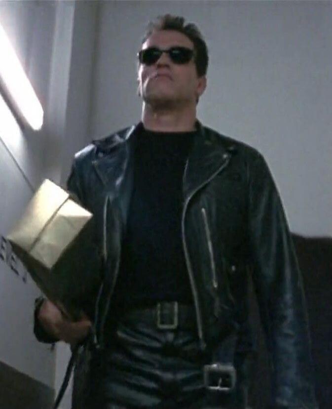 Arnold Schwarzenegger as the Terminator, wearing a black leather jacket and sunglasses, carrying a golden box. Text 'EVE J' is partially visible on the wall.