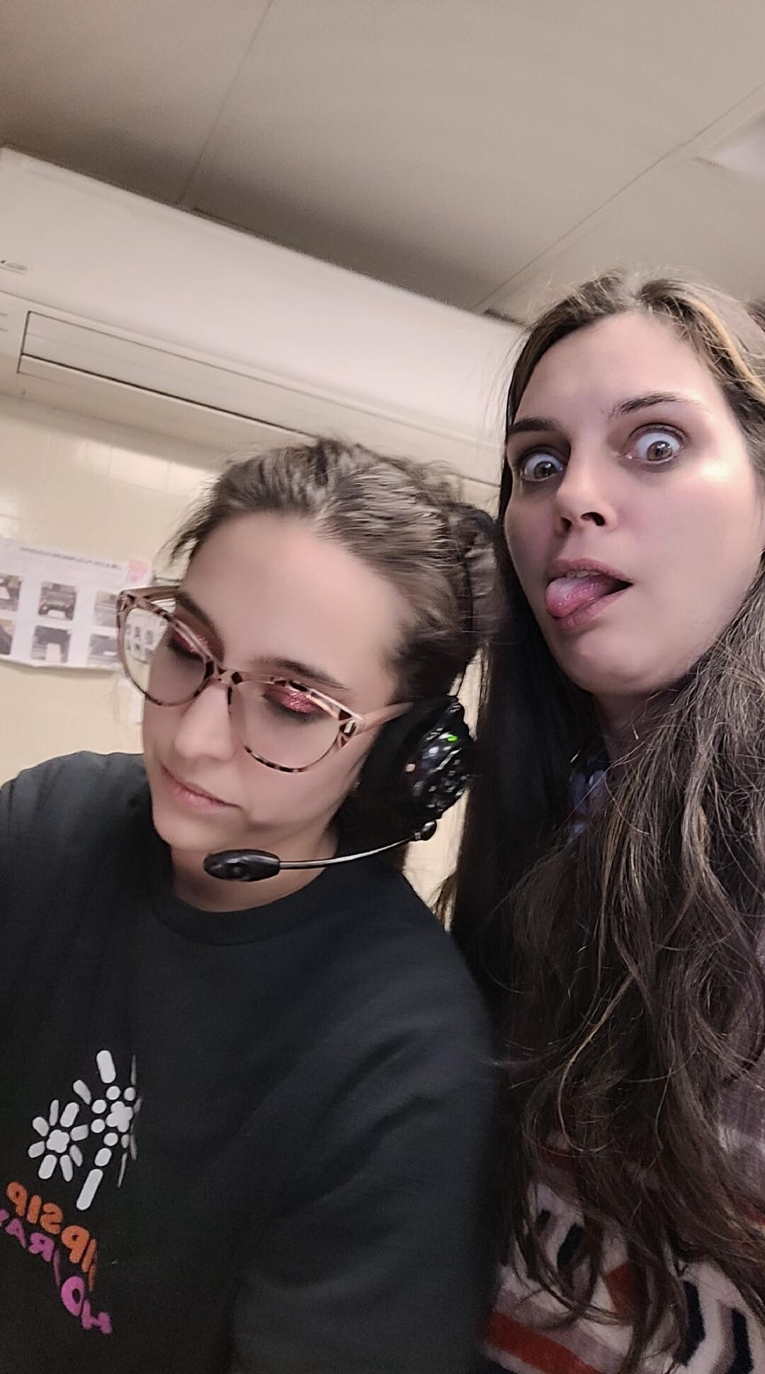 Two women are in the frame. One woman on the left is wearing glasses and a headset with a microphone, looking down, seemingly focused on something. She has glittery pink eyeshadow. Her t-shirt has some text, but it's mirrored and hard to read. The other woman on the right is making a funny face, sticking out her tongue and widening her eyes. The ba