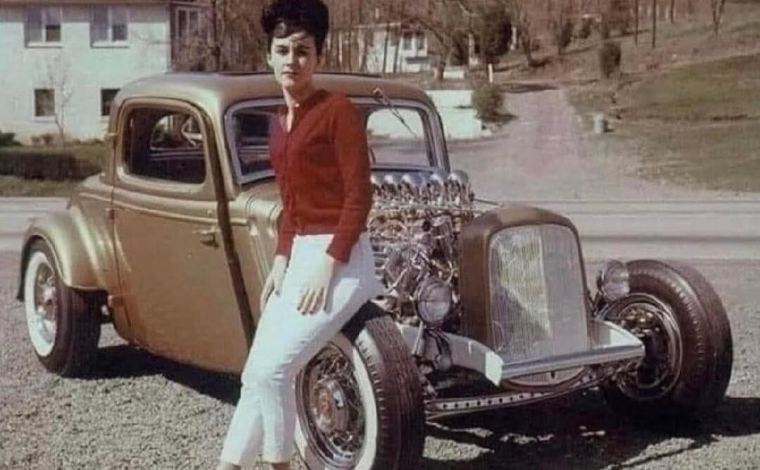 A vintage photograph of a woman in a red cardigan and white pants posing next to a custom gold hot rod car with an exposed engine. The car has large wheels with white sidewalls. A house and a road are visible in the background.