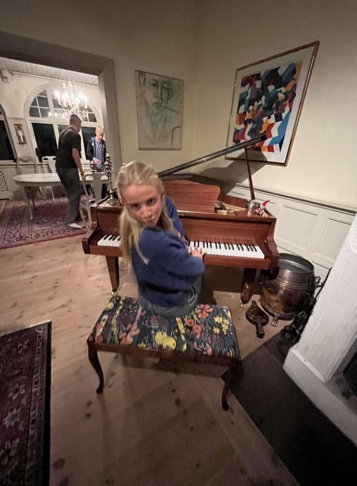 A young blonde girl is sitting on a piano bench, looking directly at the camera. She is wearing a blue sweater and jeans. Behind her is a grand piano. In the background, two men are visible in an adjacent room with a chandelier.