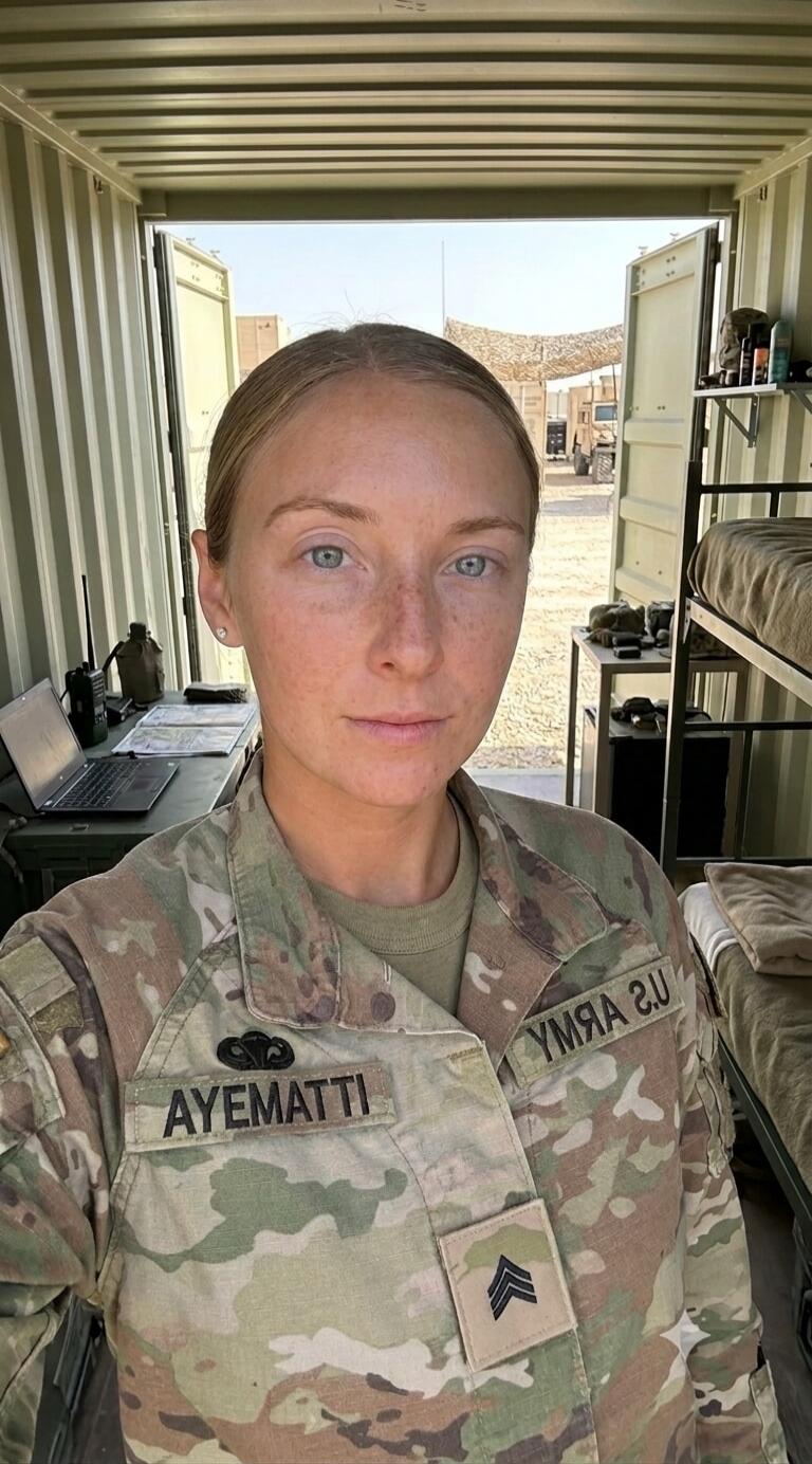 A woman in a U.S. Army uniform, with a name patch 'AYEMATTI' and a Sergeant rank insignia, takes a selfie indoors. She has light hair, blue eyes, and freckles. In the background, a desk with a laptop and a radio, as well as bunk beds, are visible. Outside, a desert environment with military structures can be seen.