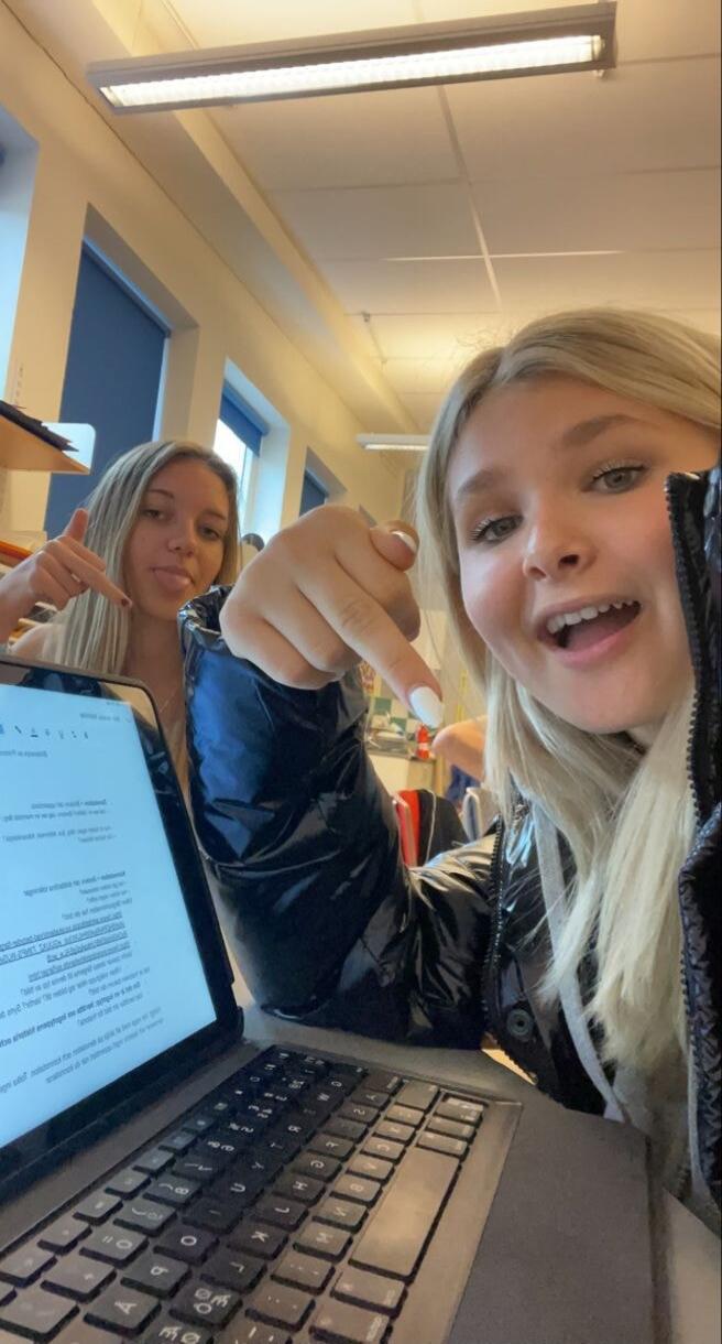 Two girls in a classroom posing for a selfie. One is pointing at the camera while the laptop on the desk displays a document.