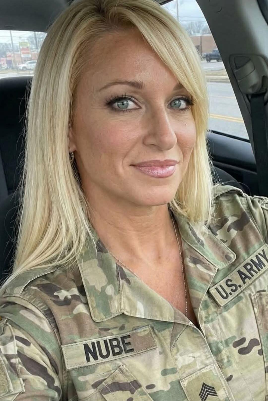 A blonde woman in a U.S. Army uniform, with a name tag 'NUBE' and 'U.S. ARMY' patch, is smiling and taking a selfie in a car. Her uniform is slightly open at the collar, revealing a necklace.