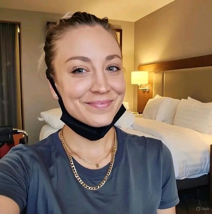 A woman smiling in a hotel room with a face mask pulled down under her chin. She is wearing a dark t-shirt and two gold chain necklaces. A bed with white linens is visible in the background.