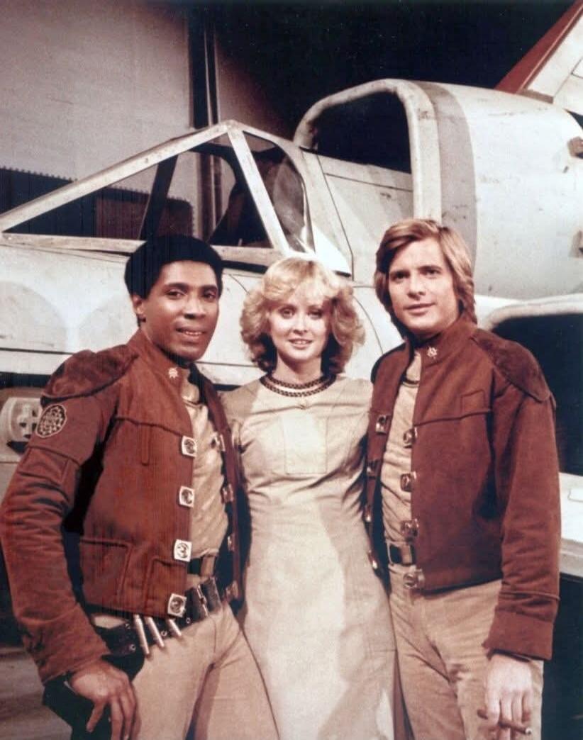 Three actors in sci-fi costumes posing in front of a spaceship, likely from the vintage television series 'Buck Rogers in the 25th Century'. The cast members are Gil Gerard, Erin Gray, and Thom Christopher.