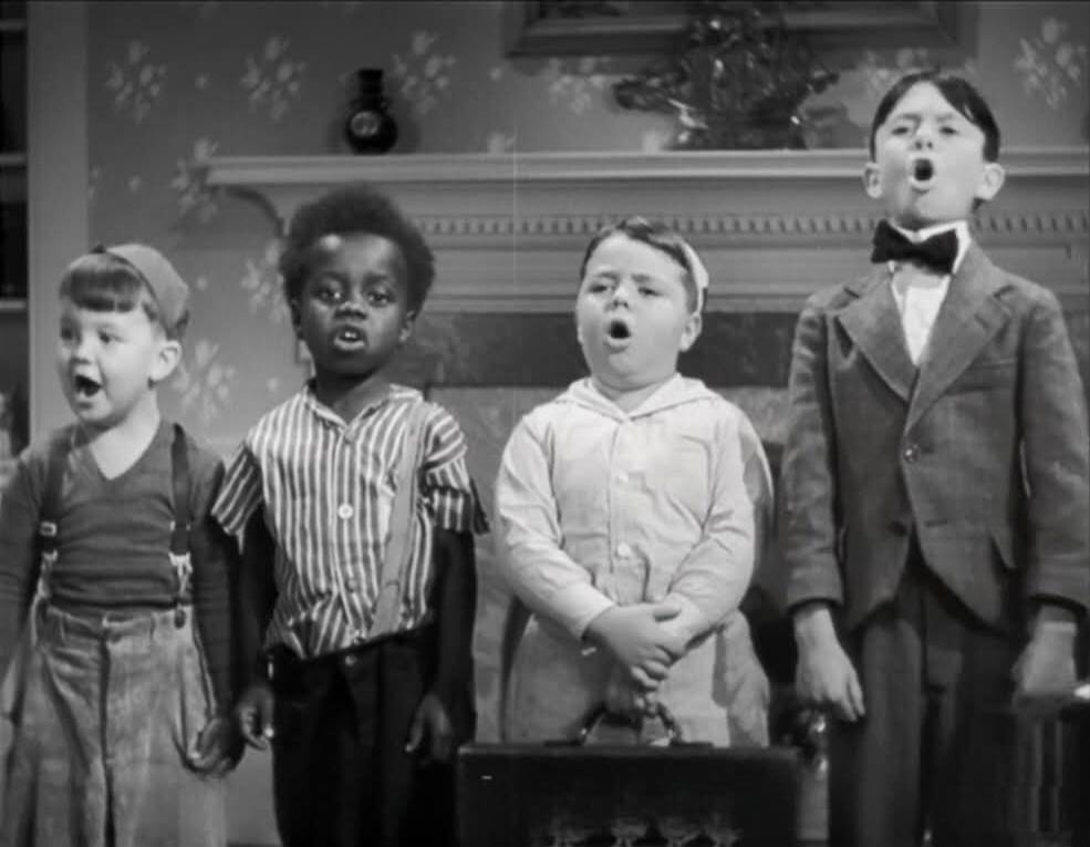 Four young boys are standing in a line with their mouths open, appearing to sing or shout.