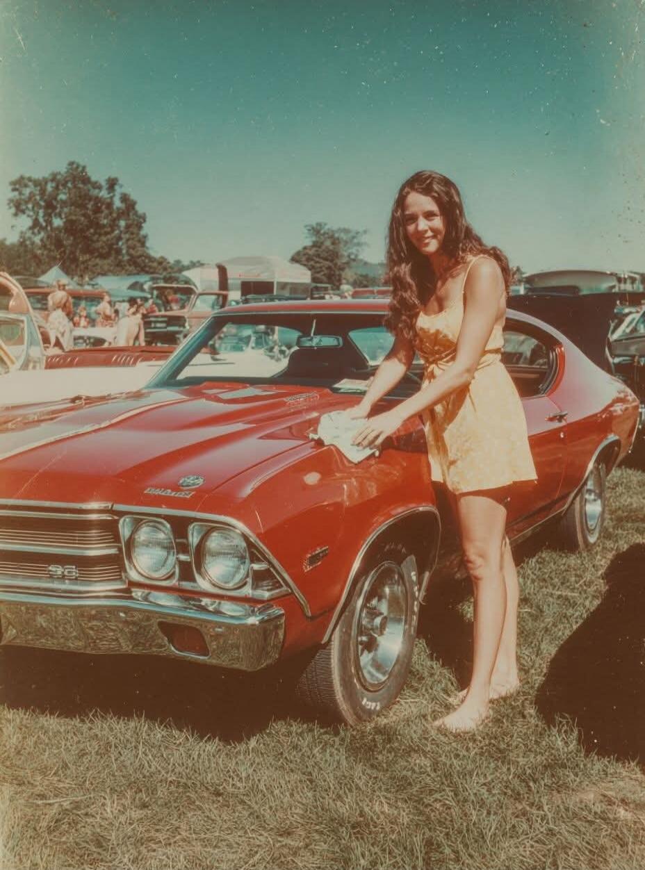 A woman in a yellow dress is wiping the hood of a red classic Chevrolet Chevelle SS car with a white cloth. She is barefoot and smiling. The scene takes place outdoors at what appears to be a car show or gathering, with other vehicles and people in the background. The car has 'SS' badging on the grille.