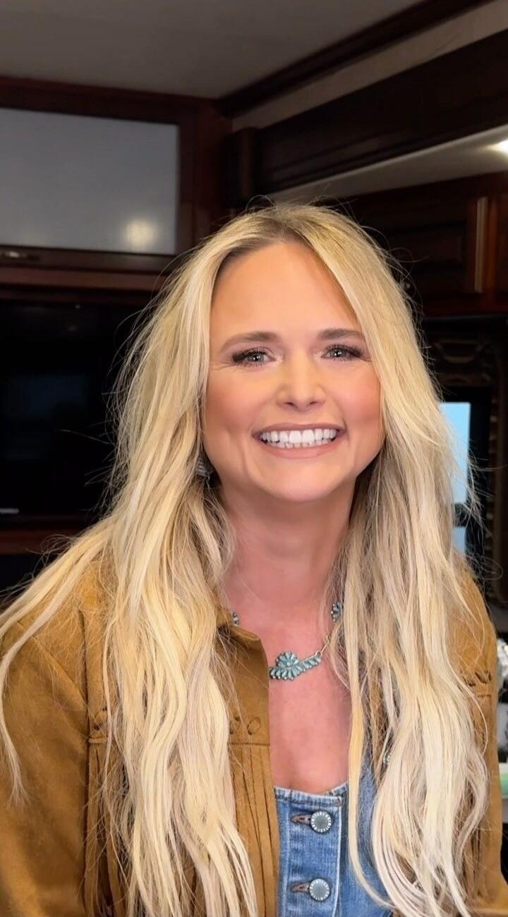 A blonde woman with long hair is smiling brightly at the camera, wearing a brown fringed jacket and a turquoise necklace.