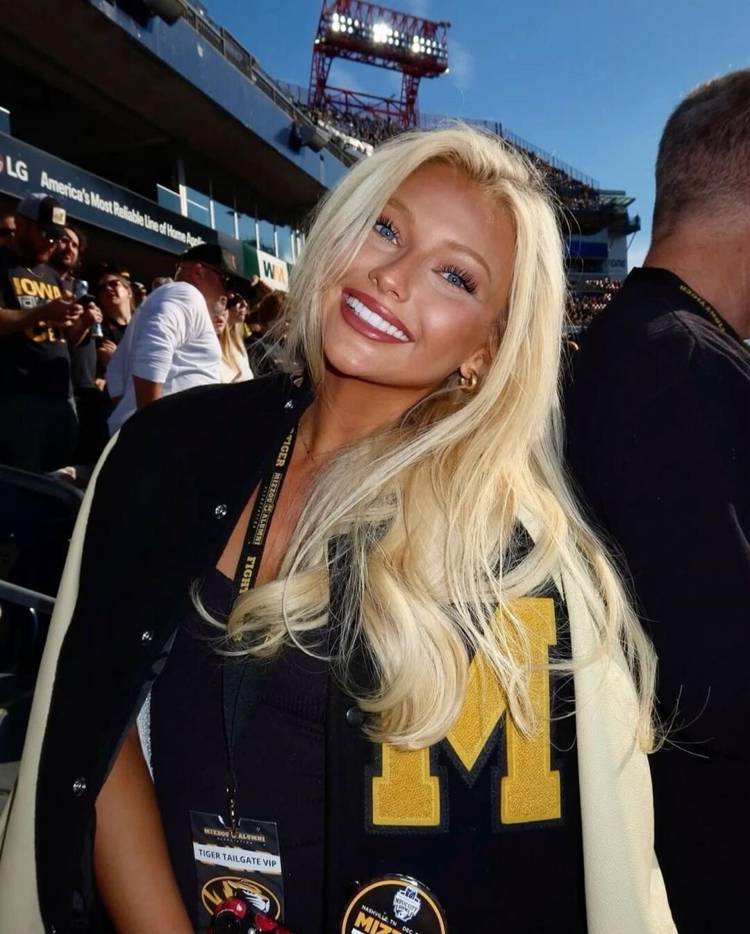 A smiling blonde woman at a stadium wearing a black jacket with a large yellow 'M' on it. She has a lanyard with a badge around her neck.