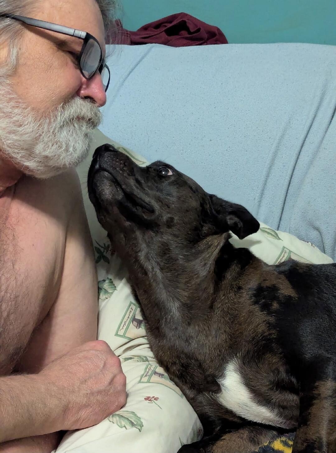 A man with a white beard and glasses is looking affectionately at a dark-furred dog. The dog is looking up at the man, appearing to share a tender moment.