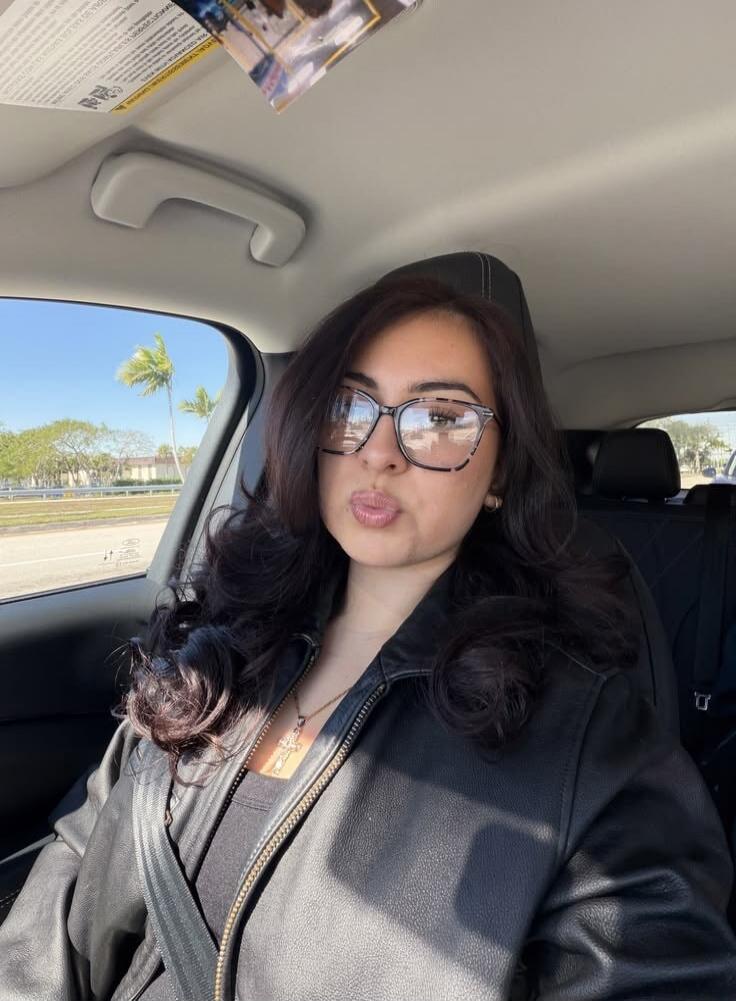 A young woman with dark hair and glasses is sitting in a car, wearing a black leather jacket and a seatbelt, making a puckered lip expression. Palm trees are visible outside the window.