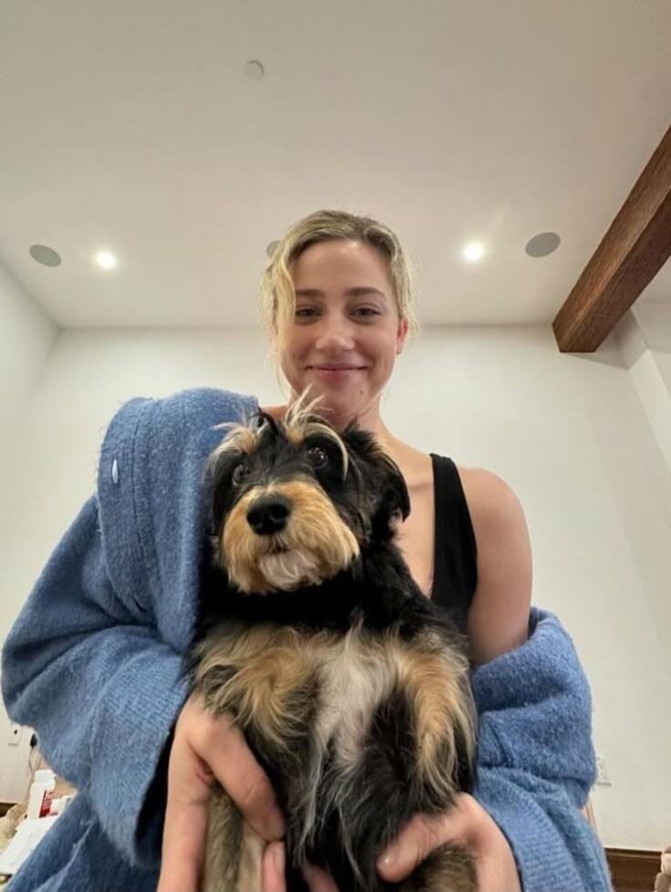 A woman is holding a small, fluffy dog and smiling at the camera. She is wearing a black tank top and a blue robe.