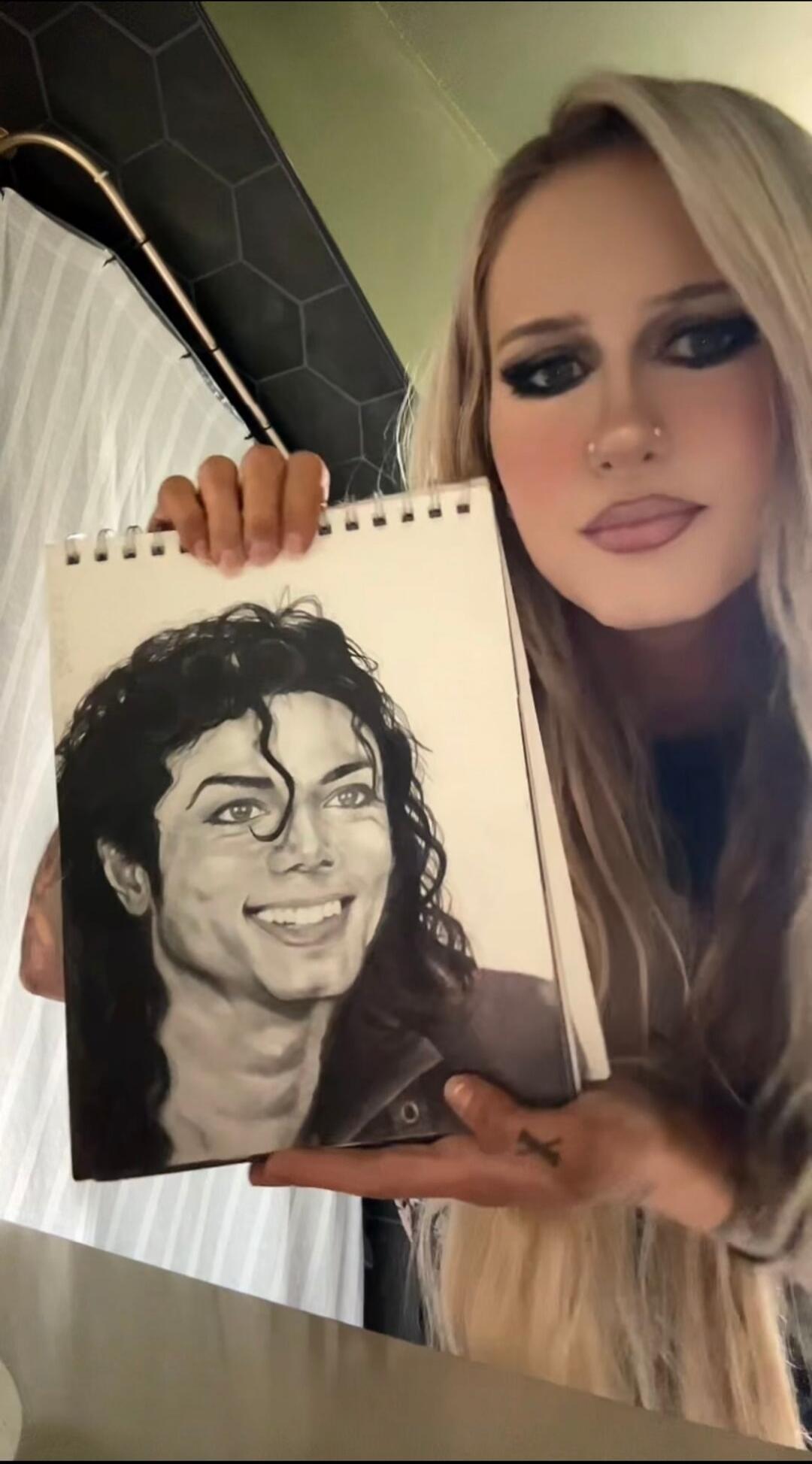 A woman with long blonde hair holds up a sketchbook displaying a black-and-white portrait of Michael Jackson.
