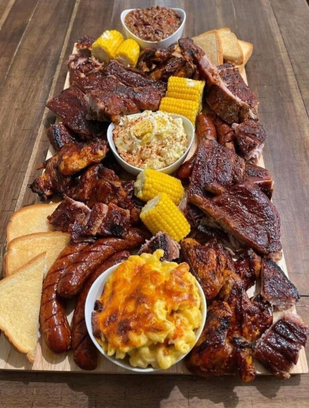 A large BBQ meat platter featuring ribs, sausages, corn on the cob, bread, and bowls of dips/sides.