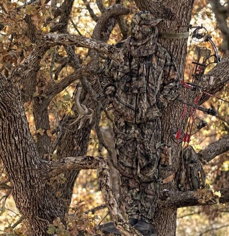A person in full camouflage hunting gear is standing on a tree stand, perfectly blending with the tree and its surroundings. A bow and arrow are visible, along with deer antlers hanging from a branch nearby.