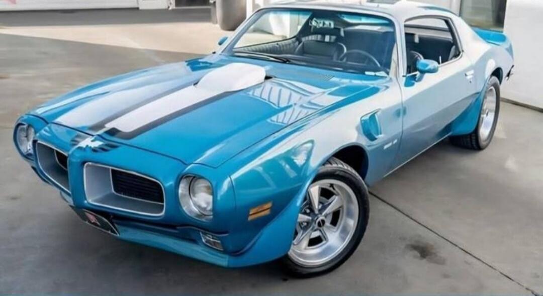A classic blue Pontiac Firebird Trans Am with white racing stripes on the hood, parked on a concrete surface.