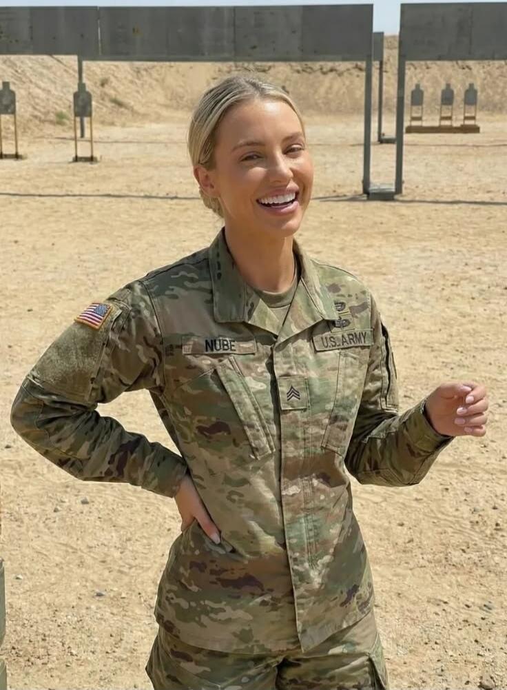 A woman in a US Army uniform with a Staff Sergeant rank insignia and an American flag patch. The name tag reads 'NUBE'. She is smiling at what appears to be a shooting range with targets in the background.