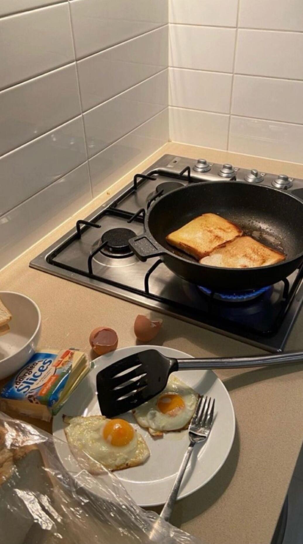 Two sunny-side-up eggs on a plate with forks and spatula, pan frying bread slices on a stove, butter packets and eggshells on counter.