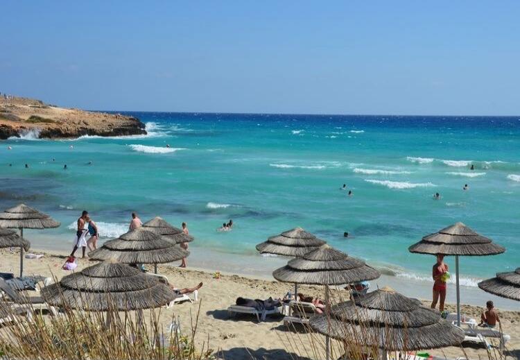 A beautiful sunny day at the beach with clear blue water and people swimming and sunbathing.