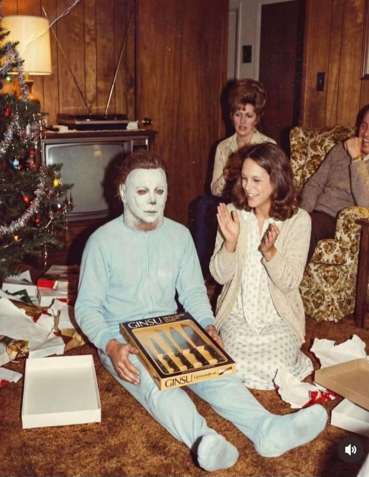 A vintage Christmas scene: a man in a white mask sits on the floor beside a boxed Ginsu knife set, while a woman in a cardigan sits nearby clapping. A Christmas tree with presents is visible, and a retro living room setting with wood-paneled walls.
