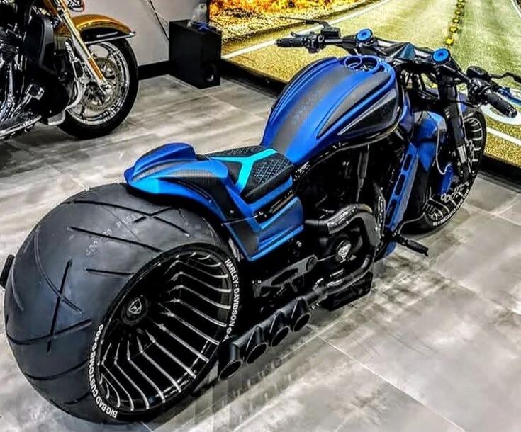 This is a custom blue Harley-Davidson motorcycle with a very wide rear tire and a unique wheel design. The motorcycle has a sleek, aggressive look with black accents and custom exhaust pipes.