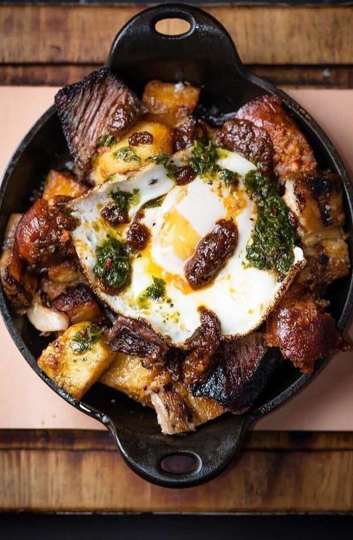 A skillet dish featuring roasted meat chunks, potatoes, and a central fried egg topped with herbs (likely parsley) and some crispy bits.