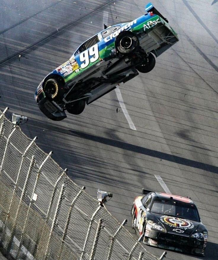 Car number 99, sponsored by Clarion and Aflac, is airborne and upside down during a high-speed race. Another car, number 09, is visible on the track below, near a safety fence.