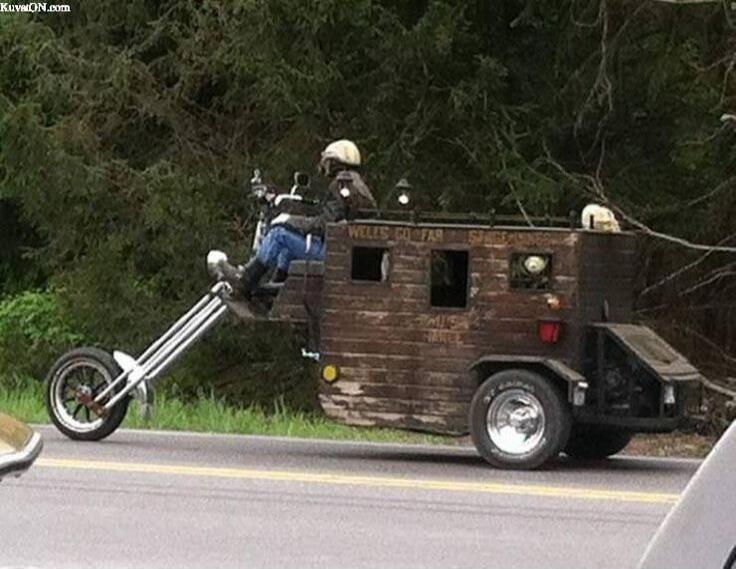 A person riding a highly customized vehicle, which appears to be a motorcycle with an extended front fork combined with a wooden stagecoach-like rear section. The side of the wooden part has text that seems to be a parody of 'Wells Fargo', reading 'WELLS GO FAR'.