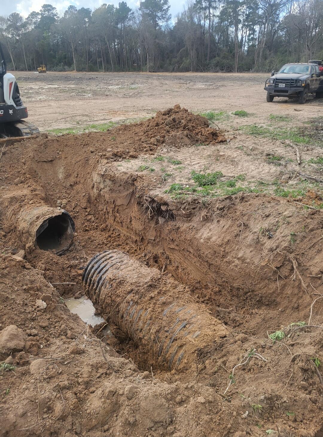 A construction site with a large corrugated drainage pipe laid in a trench. Dirt piles around. A small excavator is on the left, and a black pickup truck is on the right. Trees and open ground in the background.