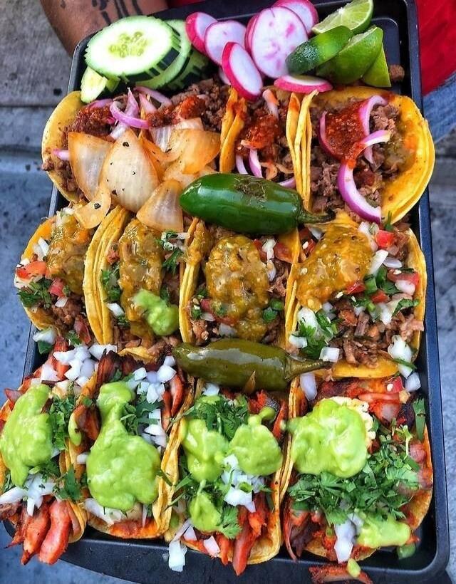Colorful tray of loaded tacos with a variety of toppings: chopped onions, cilantro, tomatoes, peppers, guacamole or avocado sauce, sour cream or crema, shredded meat, diced vegetables, lime wedges, cucumber slices, radishes, and pickled onions. A mix of crunchy toppings and fresh herbs creates a vibrant, appetizing presentation.