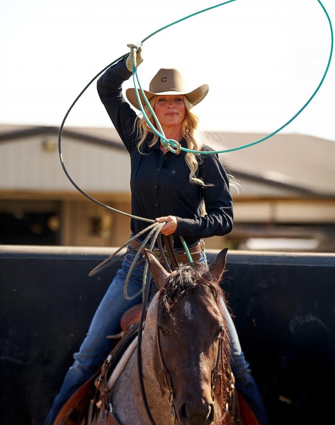A woman in a cowboy hat and black shirt, riding a horse and holding a lasso. There is a 'C' logo on her hat.