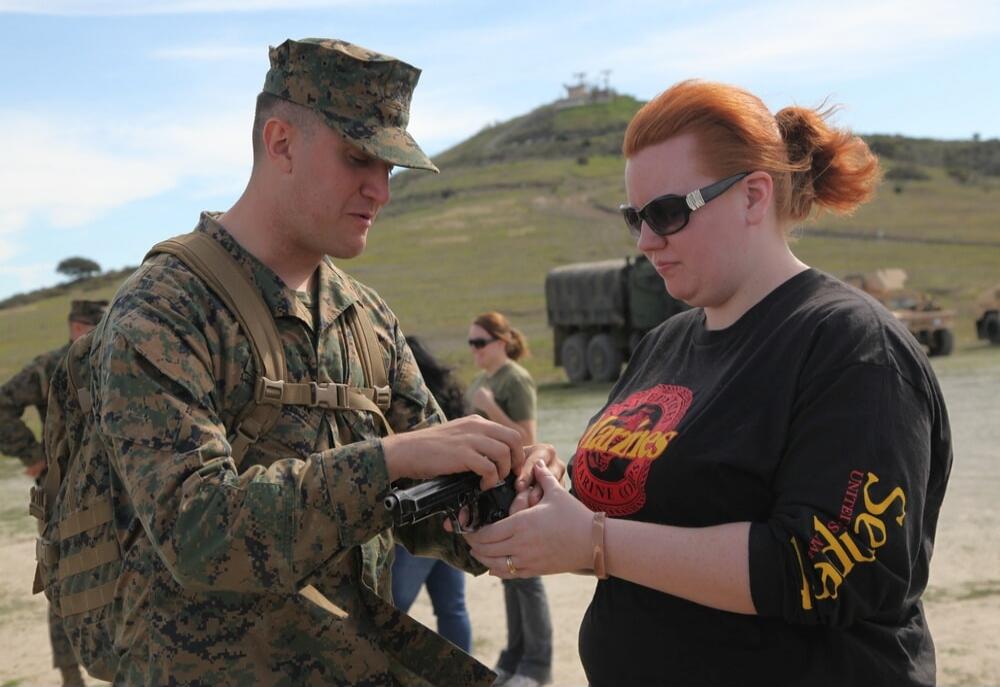 A male Marine in camouflage uniform is demonstrating how to handle a handgun to a woman wearing a black t-shirt with 'Marines' and 'United States Ladies' written on it. They are outdoors, possibly at a shooting range or a military event, with other people and military vehicles in the background.
