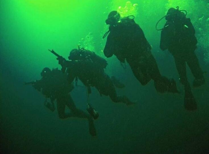 Four divers, possibly military or special forces, are seen underwater in murky green water. Two of them are holding what appear to be underwater rifles.