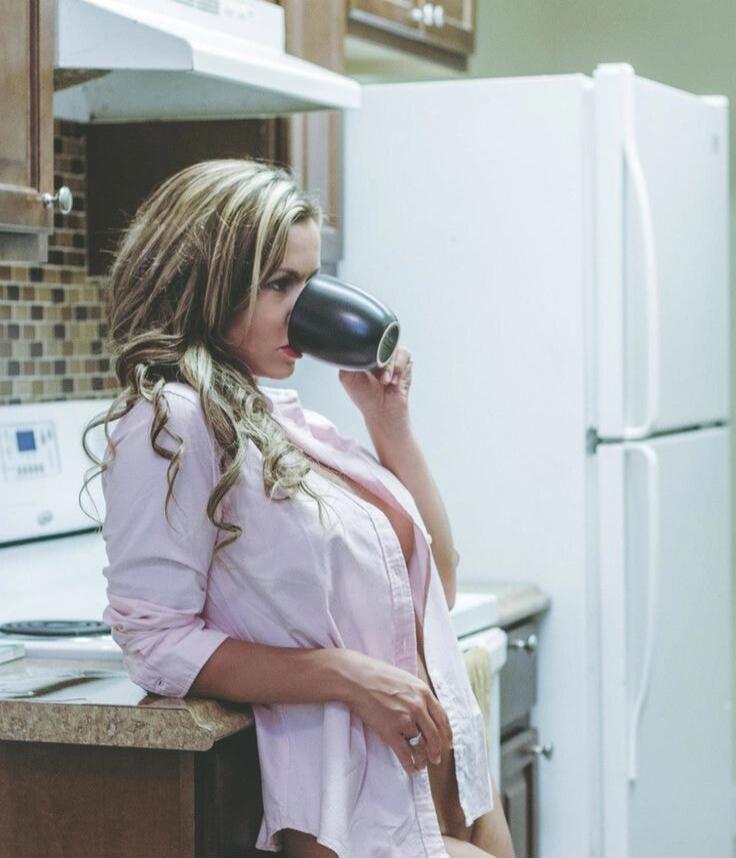 A pregnant woman in a pink shirt leans on a kitchen counter while drinking from a black mug.