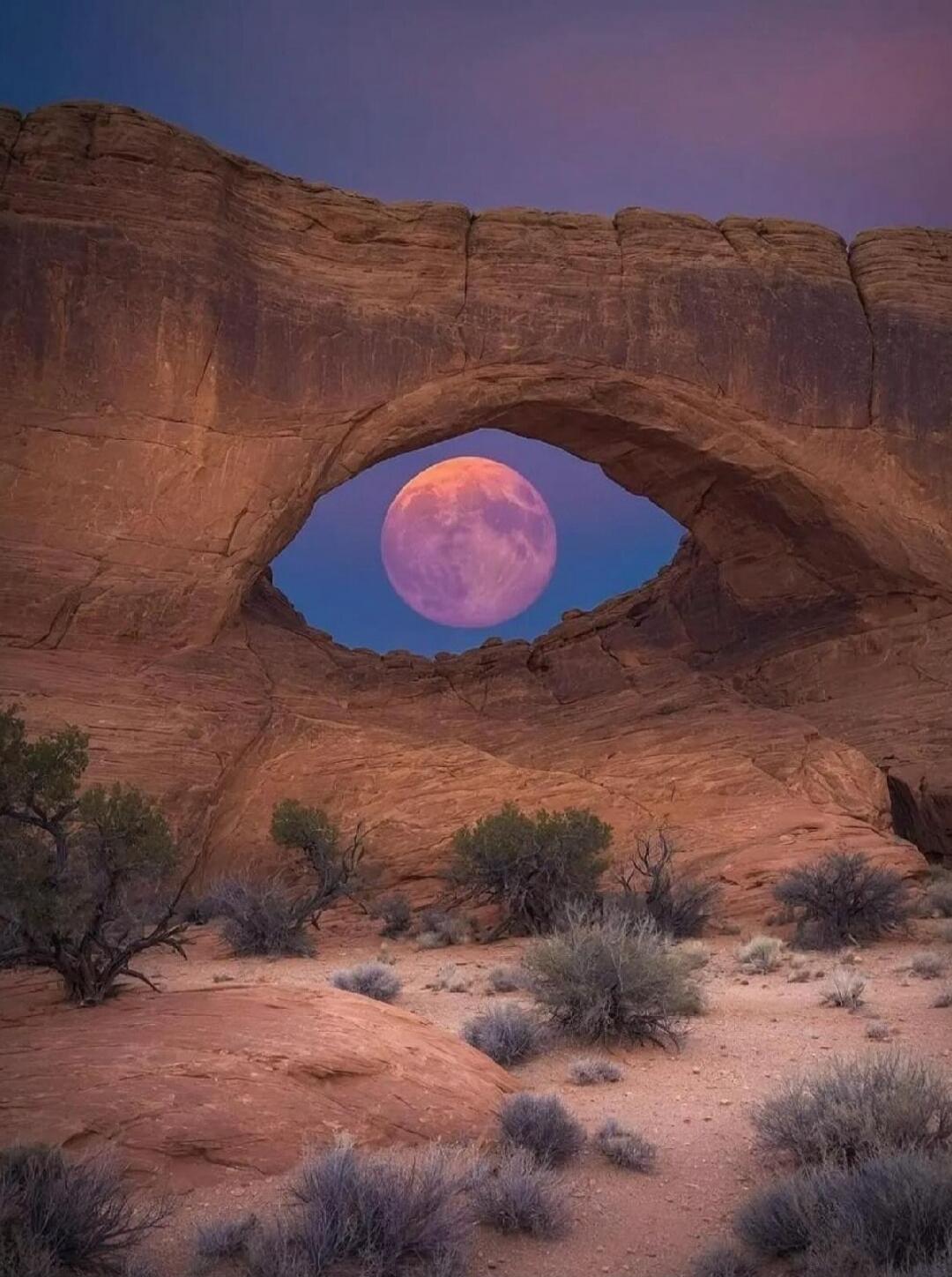 A natural rock arch with a full moon visible through the opening against a purple-tinged sky.