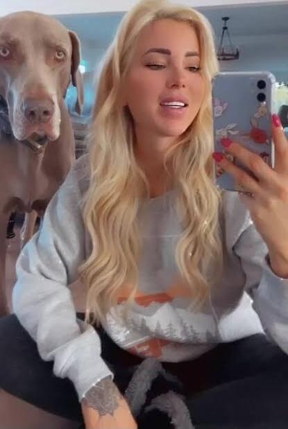 A blonde woman is taking a selfie with a large dog indoors. The dog is standing close beside her, looking at the camera.