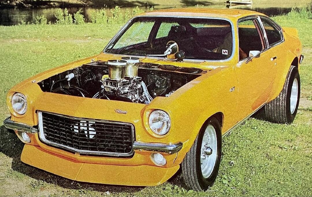 A classic yellow Chevrolet Vega GT with an open hood, showcasing a powerful modified engine with dual carburetors. The car features an SS emblem on the grille and wide rear tires, suggesting a performance build. It is parked on a grassy area next to a body of water.