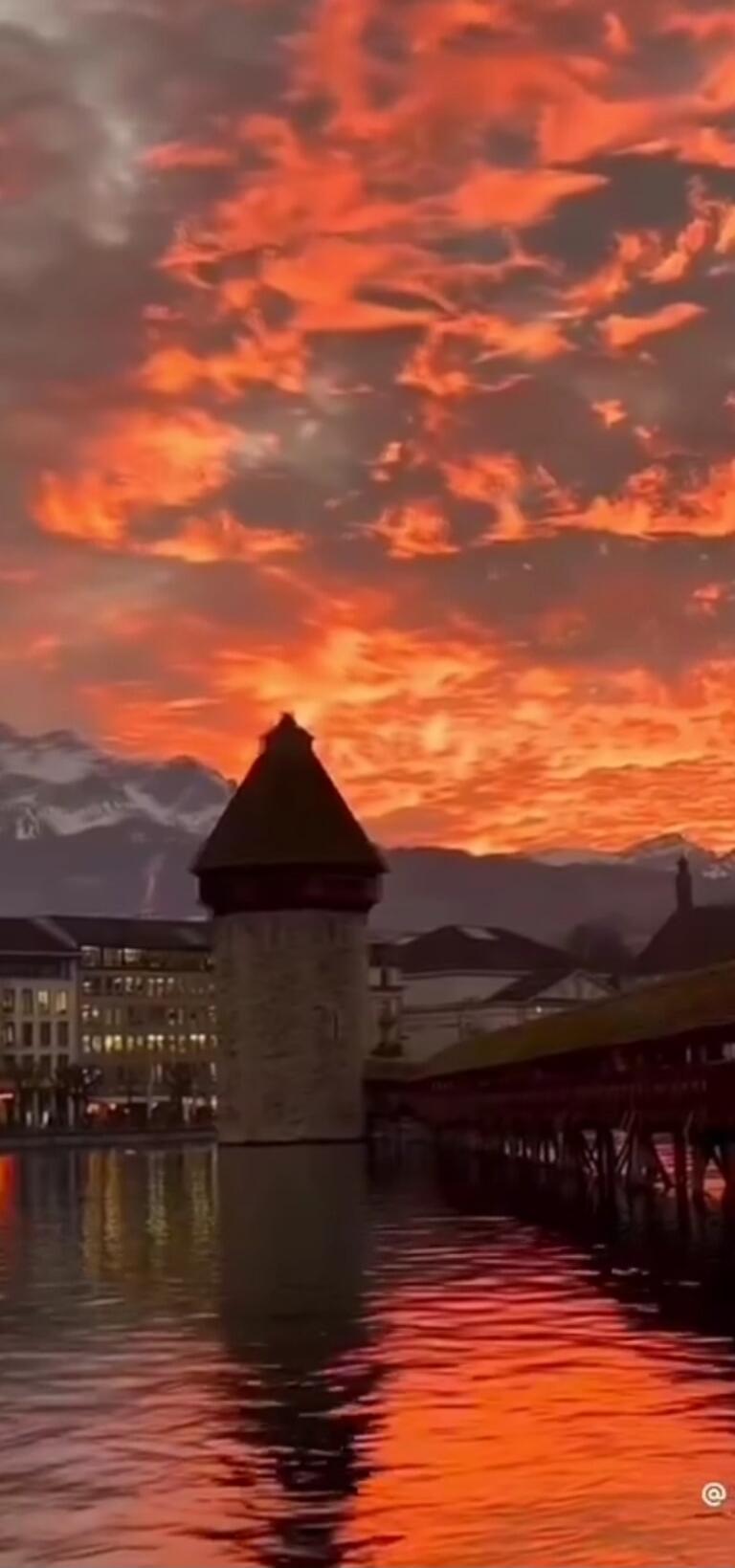 A dramatic orange sunset sky over a lake, with a stone tower standing in the water near a wooden bridge.