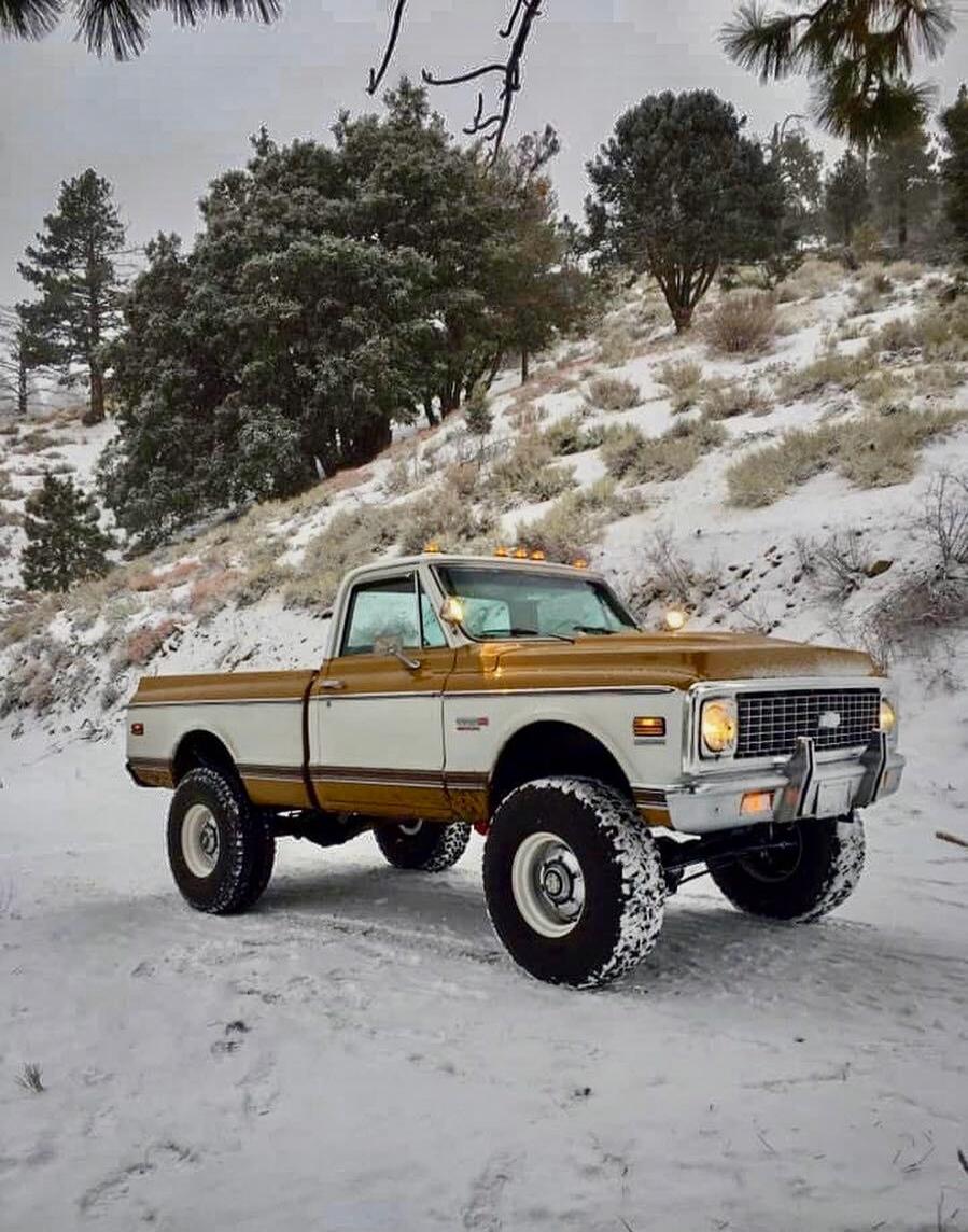 A vintage brown and white pickup truck with large tires parked on a snow-covered hillside, trees in the background.