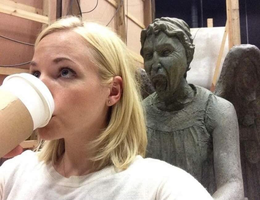 A blonde woman is drinking from a paper cup, seemingly unaware of the terrifying Weeping Angel statue from Doctor Who positioned right behind her, with its mouth open in a scream.