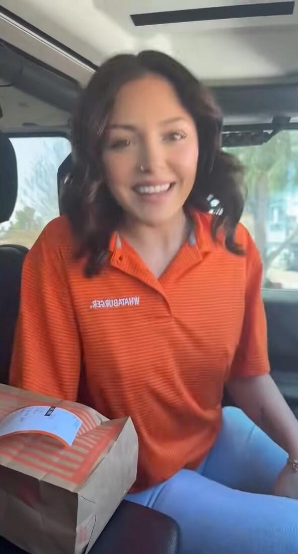 A young woman smiling in a car, wearing an orange polo shirt with 'WHATABURGER' written on it. A paper bag, likely containing food, is next to her.