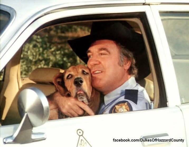 A man in a sheriff's uniform and a cowboy hat is sitting in a car, affectionately hugging a brown dog. The dog has its tongue slightly out. Text on the image reads: facebook.com/DukesOfHazzardCounty.
