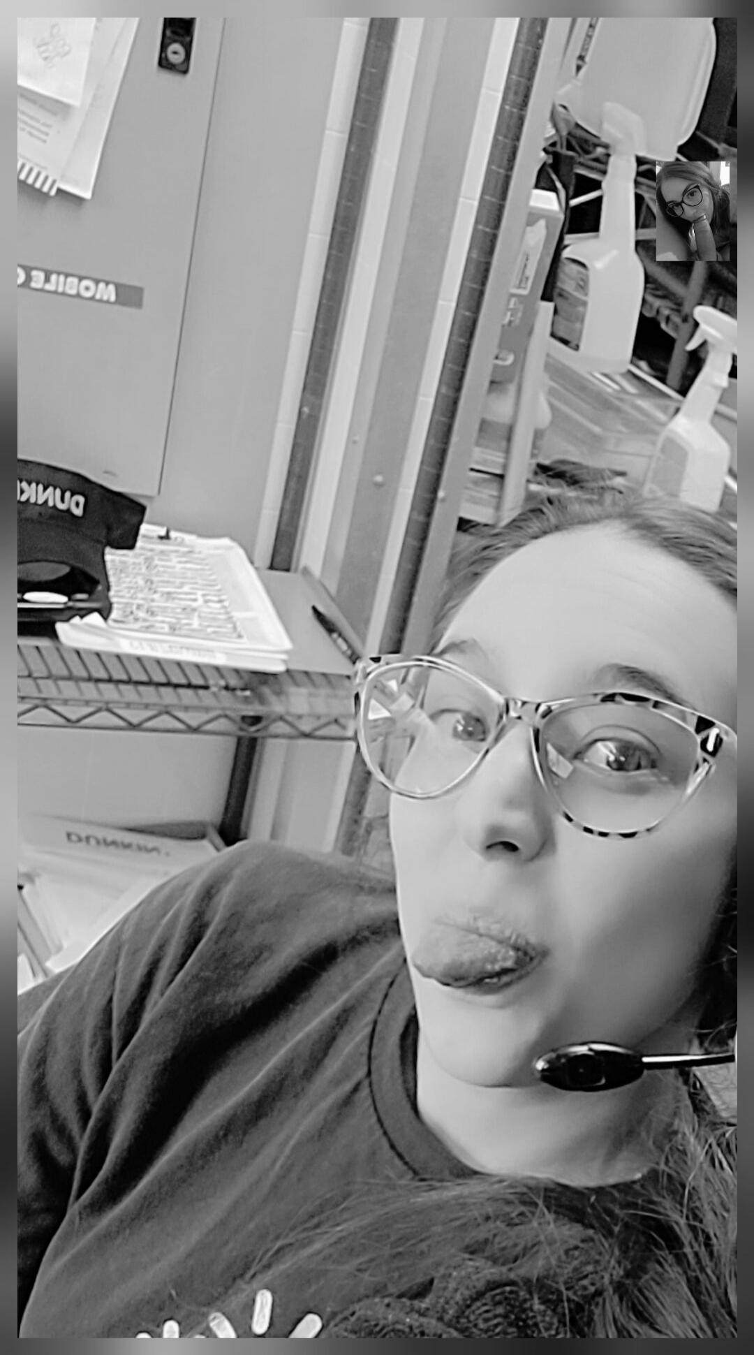A woman wearing glasses and a headset takes a selfie, sticking her tongue out playfully. In the background, reversed text like 'MOBILE' and 'DUNKIN'' is visible, suggesting a work environment.