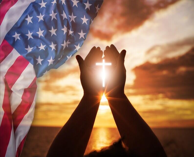 Silhouetted hands in prayer form a cross against a sunset, with the American flag in the background.