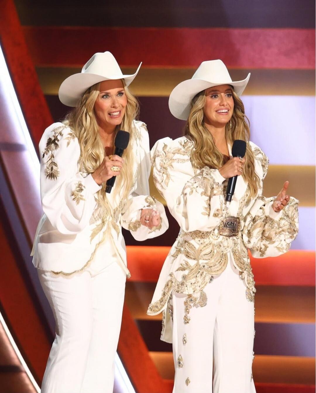 Two female performers in white outfits with cowboy hats holding microphones on stage.